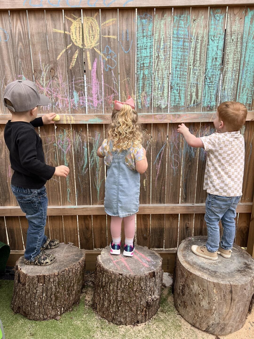 Three students standing on logs using chalk on the fence at The Nest, a preschool and learning center in Surf City, North Carolina