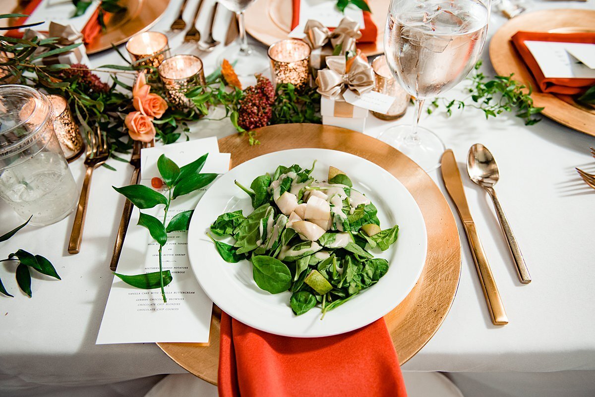 Preset salad on a white plate with a gold charger and gold flatware accented by an orange napkin