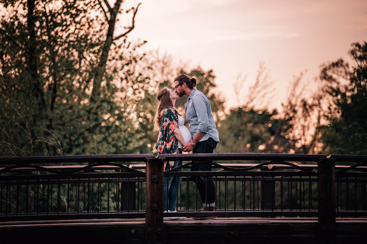 A couple expecting their first child hold hands and kiss while standing on a wooden bridge over Minnehaha Creek in Minneapolis at sunset.