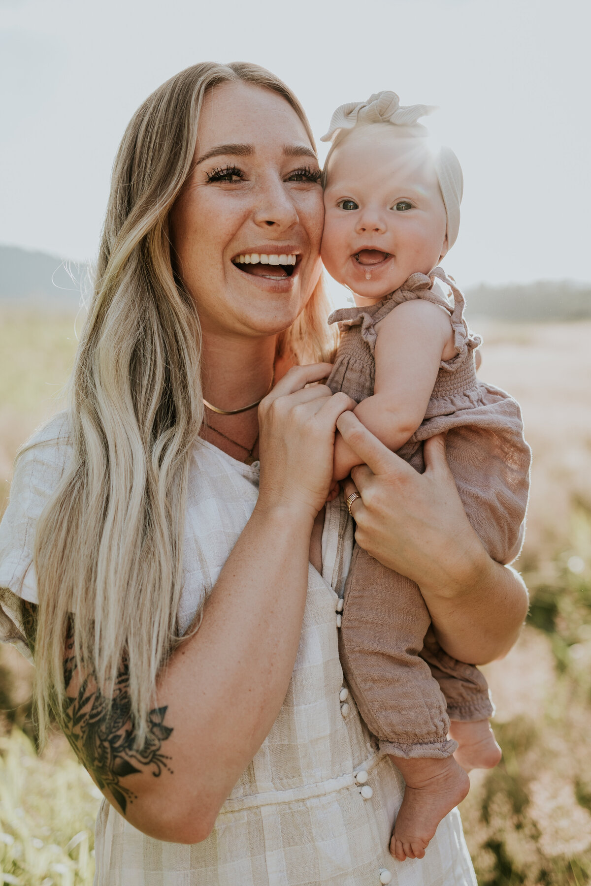 Mama snuggles smiling baby girl while wearing neutrals in the middle of a field.