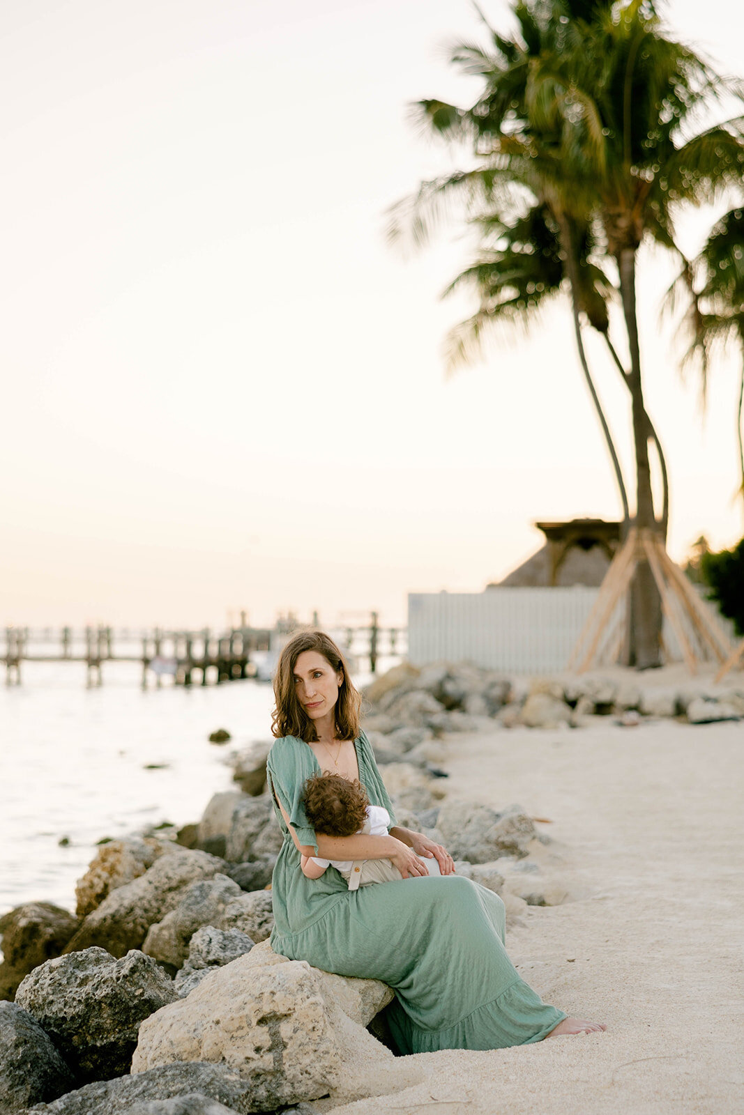 A mother in a flowing green dress sits on a rocky shore, breastfeeding her baby during a sunset Florida Keys family session. Captured by Claudia Amalia, Florida Keys family photographer.