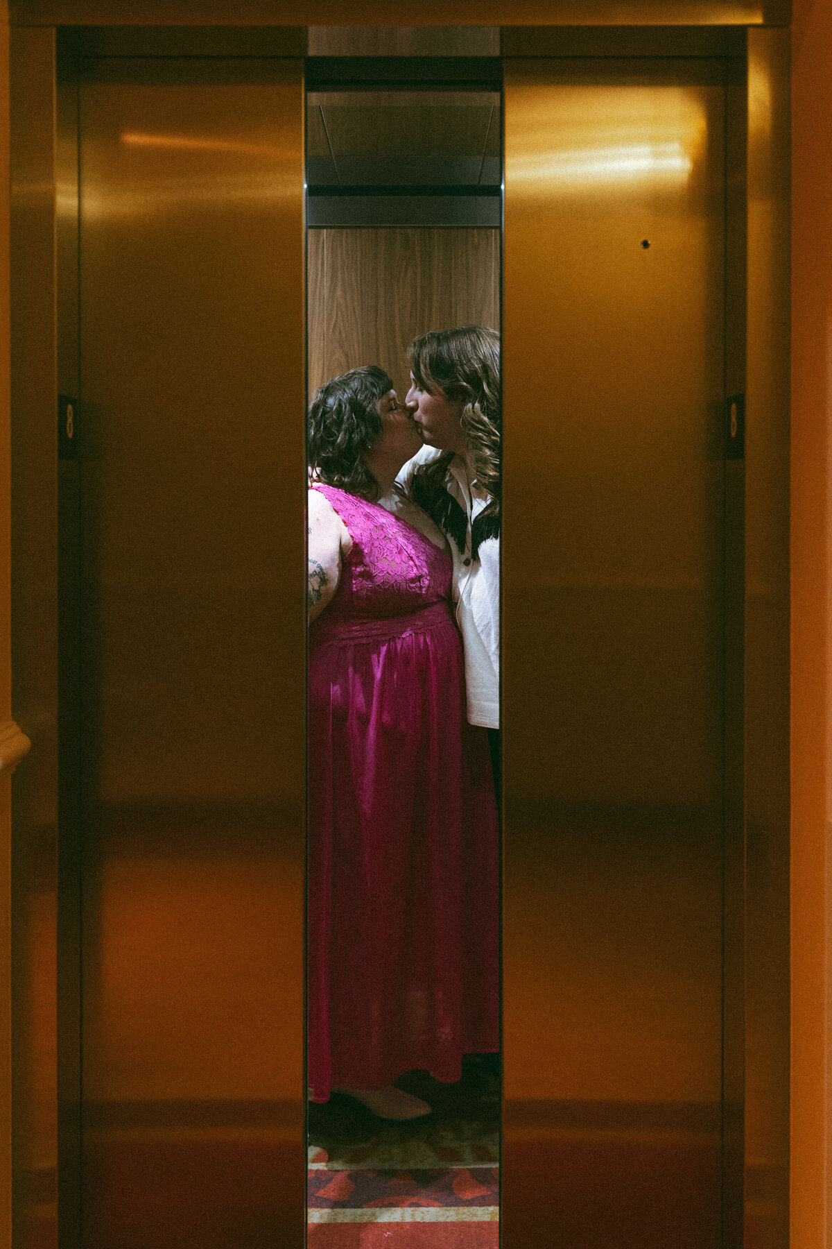 Two women sharing a moment in an elevator with warm lighting, one in a pink dress and the other in a white blouse