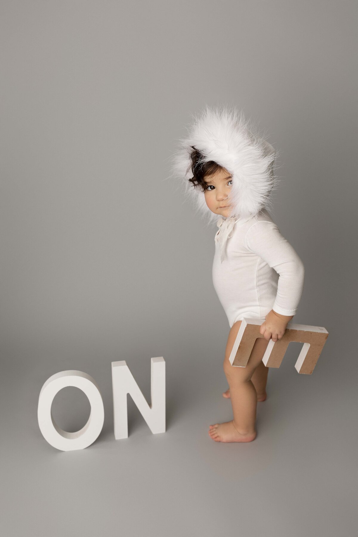 Baby in faux-fur bonnet and white onesie standing and looking at the camera as they are picking up wood o-n-e letters