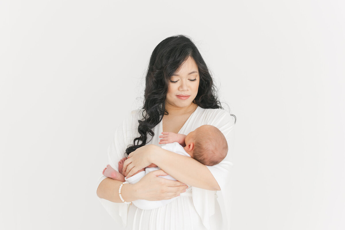 A Northern Virginia Newborn Photography photo of a mother wearing white against a white wall smiling down at her infant swaddled in a white blanket