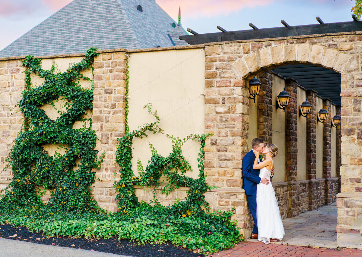 Groom leans up against the wall under an arch walway as his bride kisses him at the Pinnacle Golf Course in Grove City, Ohio. Italy inspired,  climbing vines.