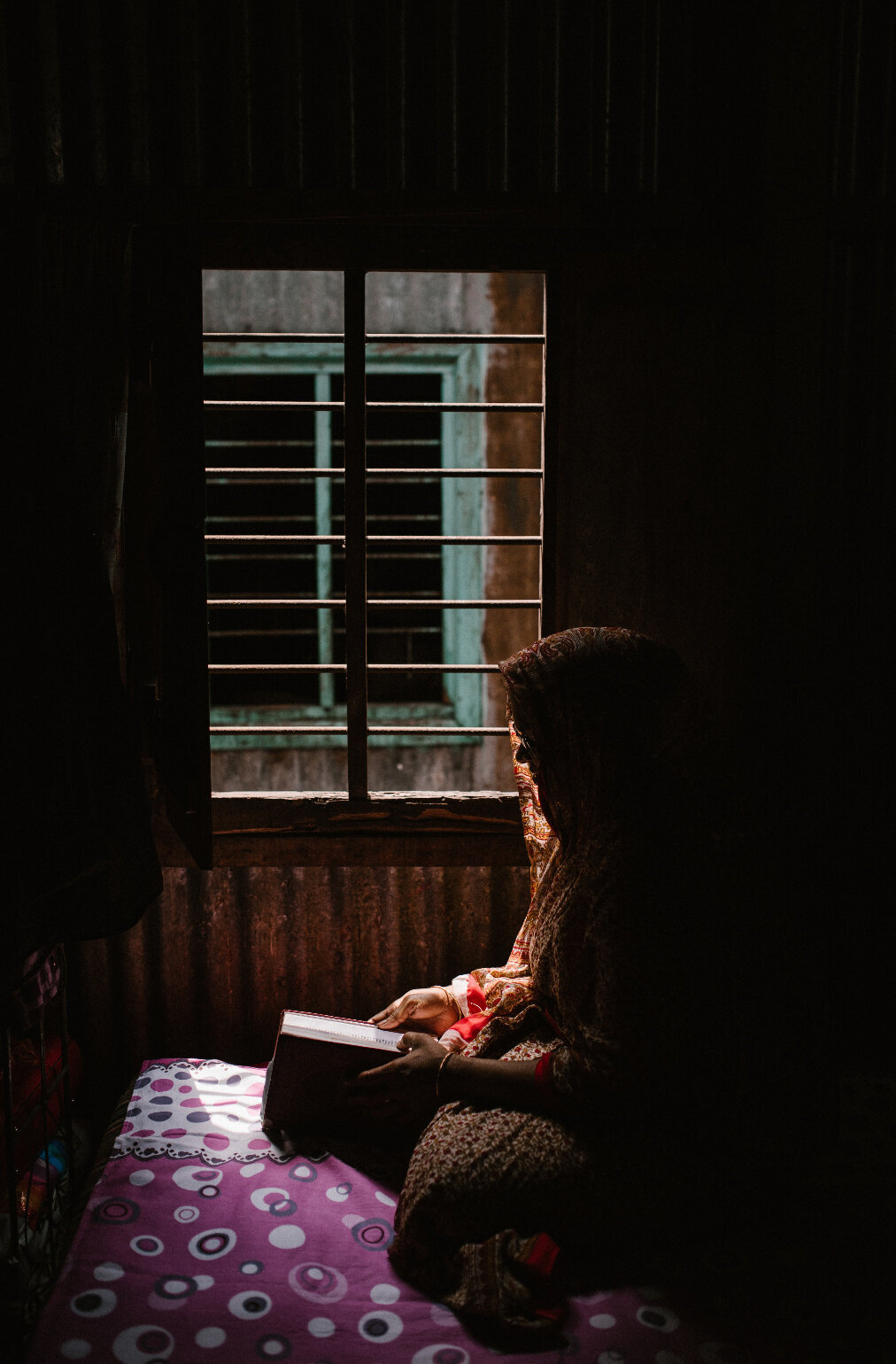 Een vrouw met een hoofddoekzit met een boek  zit voor het raam. Het licht valt naar binnen via het raam