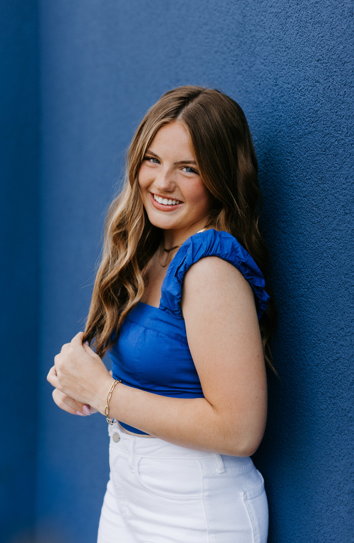 Senior girl in a blue top and white pants leaning against a blue wall