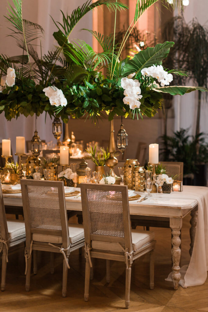 The photo was taken at the Mellon Auditorium in Washington D.C., which is known for being one of the best luxury venues in the city with European flair. Photographer Sarah Bradshaw beautifully captured a scene of a lavishly set wooden dining table with elegant tall-back chairs. Adorned with white orchids, lush greenery, lanterns, candles, and delicate dinnerware, the table exudes an air of elegance and sophistication. The ambient lighting in the room casts a warm glow, further enhancing the inviting atmosphere of the setting.