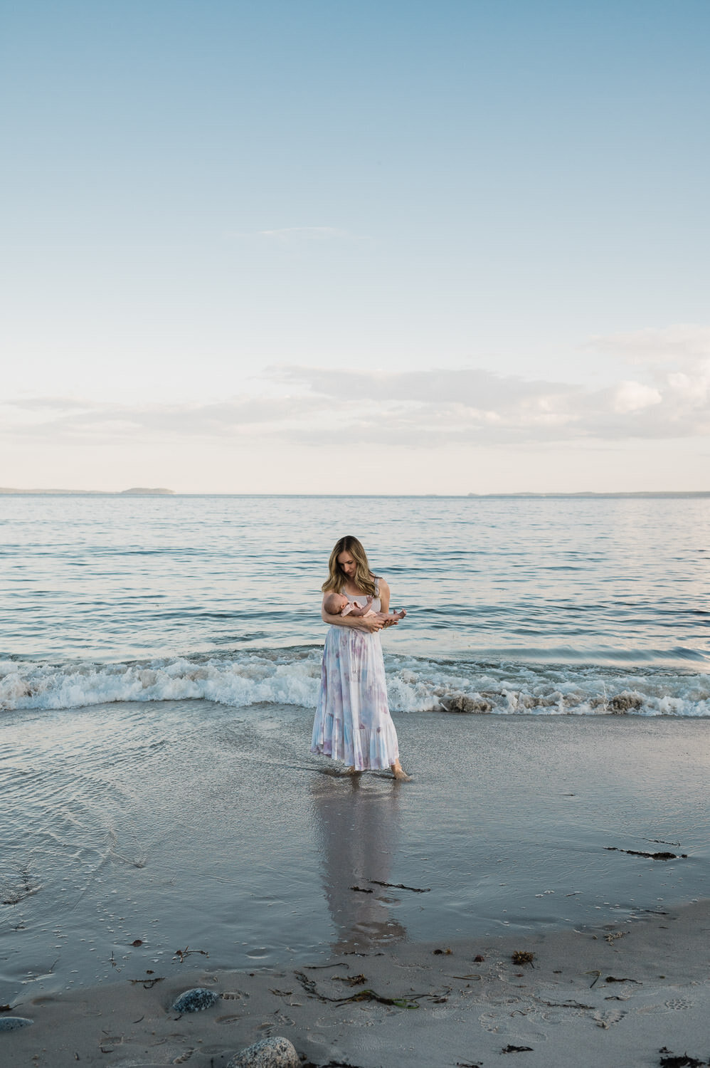 Woman holding baby while standing in water on the beach at sunset.