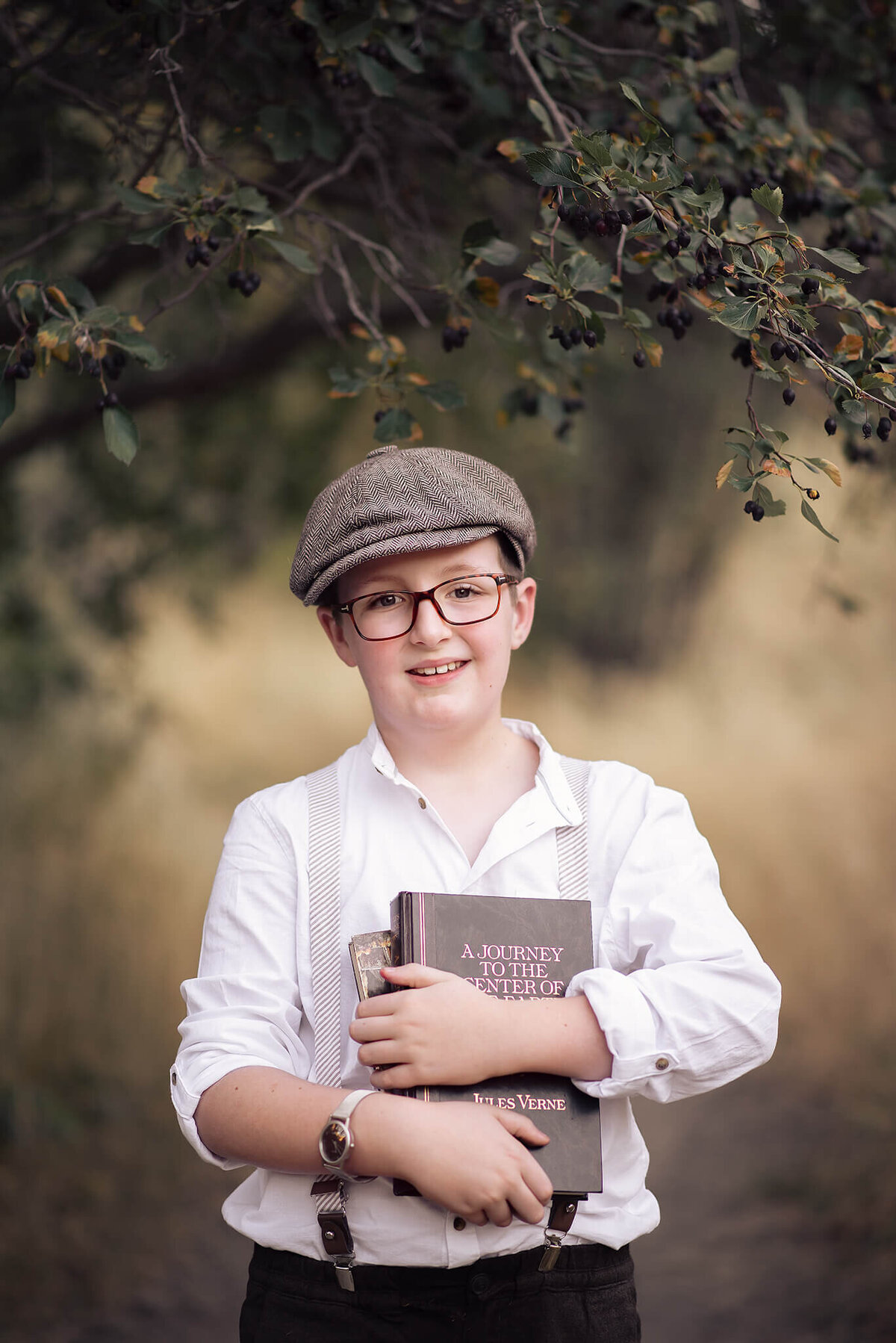 Young boy with glasses and page boy cap holding a book to his chest while smiling. Captured by Brittany Danielle Photography