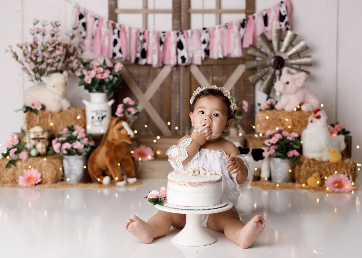Farmed theme cake smash in Wellington and Palm Beach Florida. Baby girl is wearing a lace off the shoulder romper sitting behind a white naked cake with flowers. The background is a barn inspired backdrop with barn doors, windmill, Haystacks and plush farm animals. Soft pink and cow print fabric drapes across the barn doors.