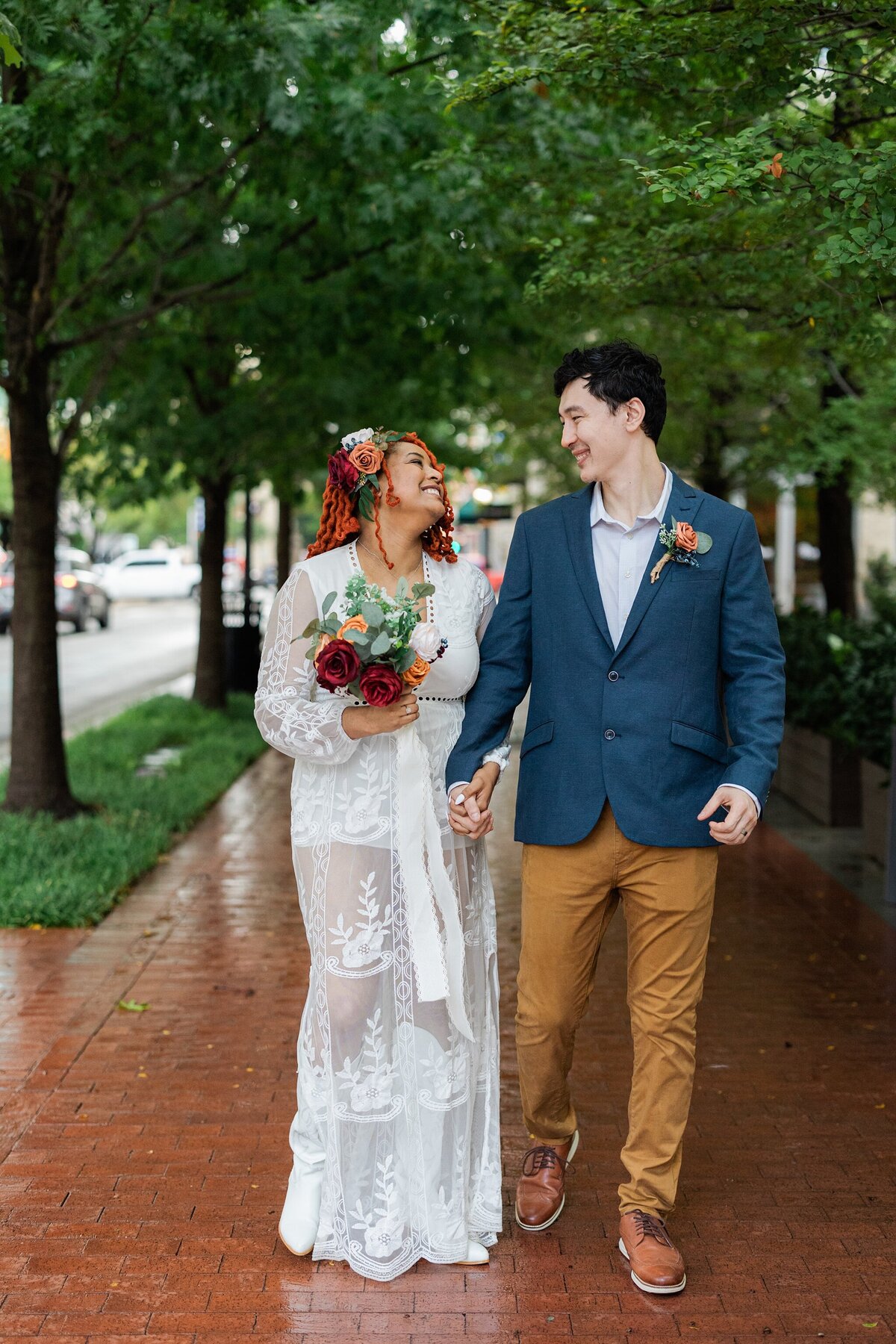 Portrait of a bride and groom walking down a brick sidewalk after their elopement at the Tarrant County Courthouse in Fort Worth, Texas. The bride is on the left and is wearing a long sleeve, intricately detailed, white dress with a floral hairpiece and is holding a bouquet. The groom is on the right and is wearing a blue jacket, tan dress pants, and a boutonniere. They hold hands and smile at each other as they walk towards the camera.