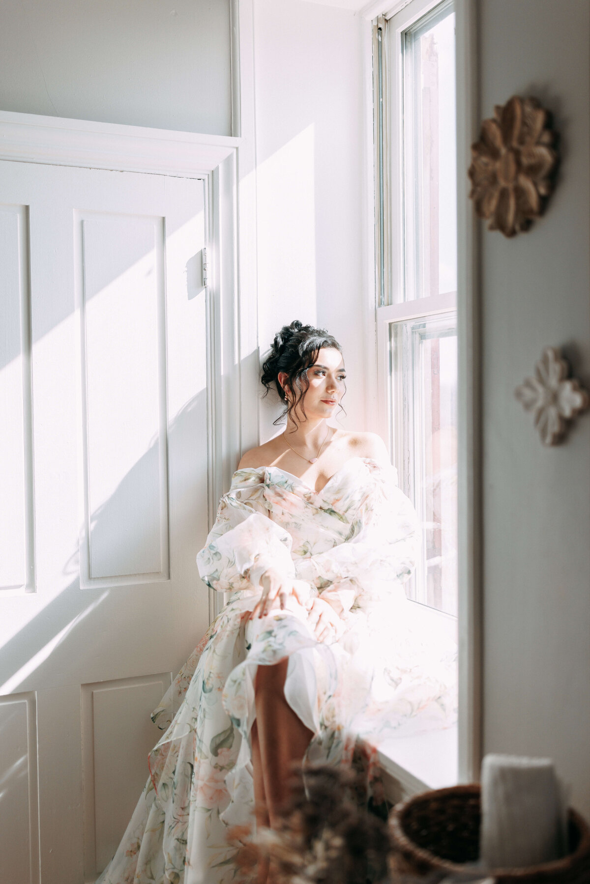 bride wearing floral wedding dress sitting by a window