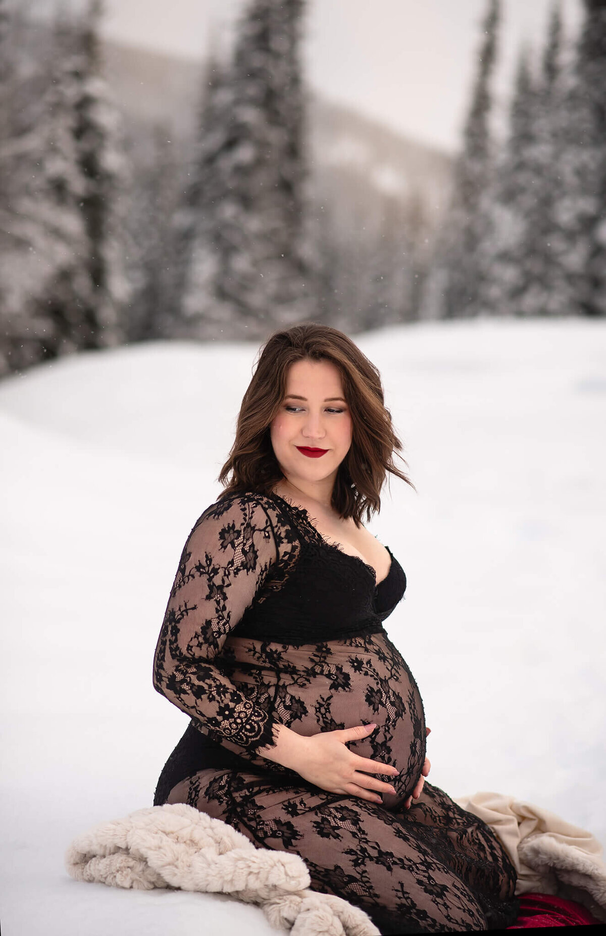 A pregnant woman with short brown hair sits on a blanket in the snow wearing a sheer black lace dress. She holds her belly while looking off and smiling. Taken by Brittany Danielle in Sun peaks