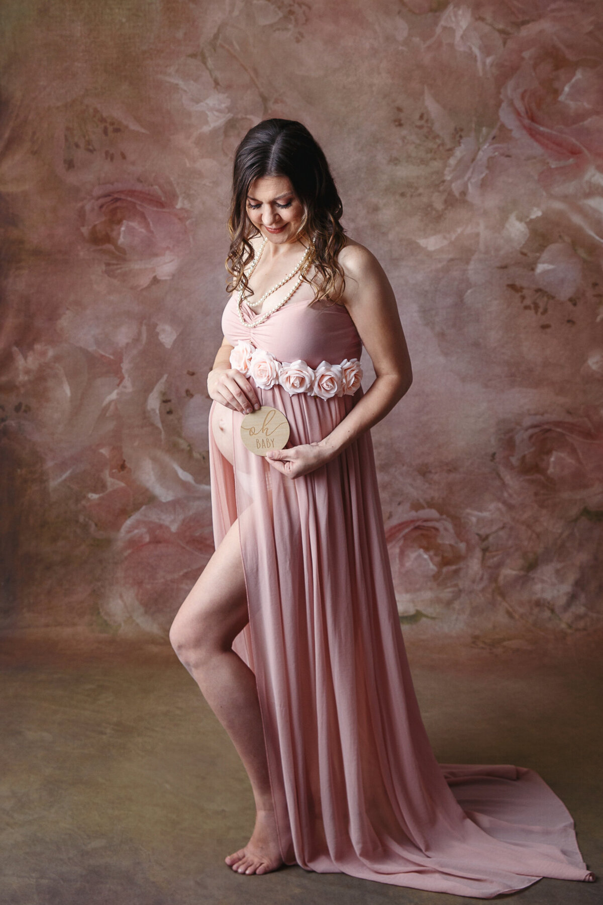 Full body portrait of a woman in a pink maternity dress holding a sign that says oh baby