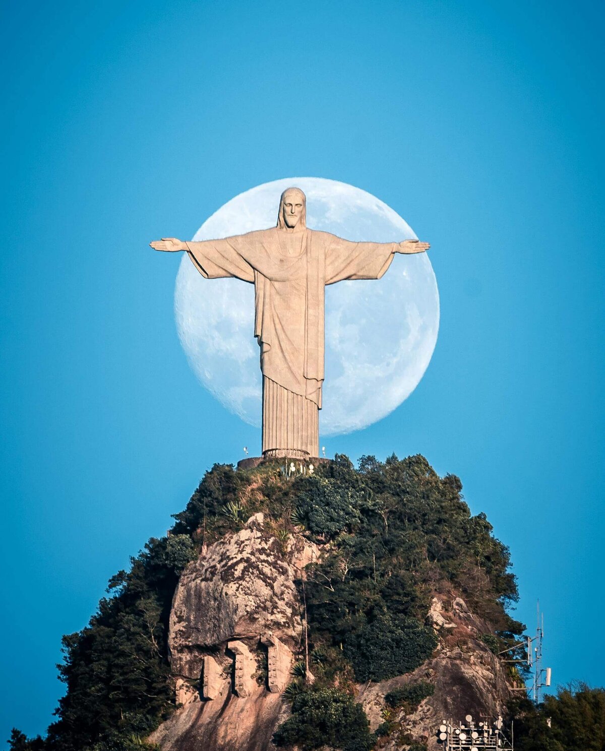 The iconic Christ the Redeemer statue atop a lush green hill, with the full moon perfectly aligned behind it against a bright blue sky. This awe-inspiring landmark reflects the grandeur of destinations supported by vacation fundraising for charities.