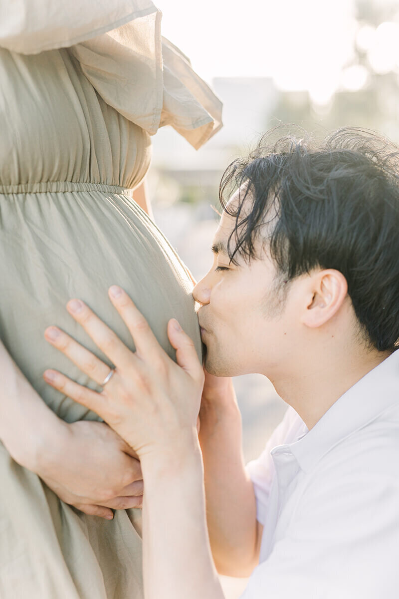 カーキベージュのワンピースを着た妊婦さんのおなかに手を添えてキスをするパパの写真。夕日の光が美しい、おしゃれなマタニティフォト。
