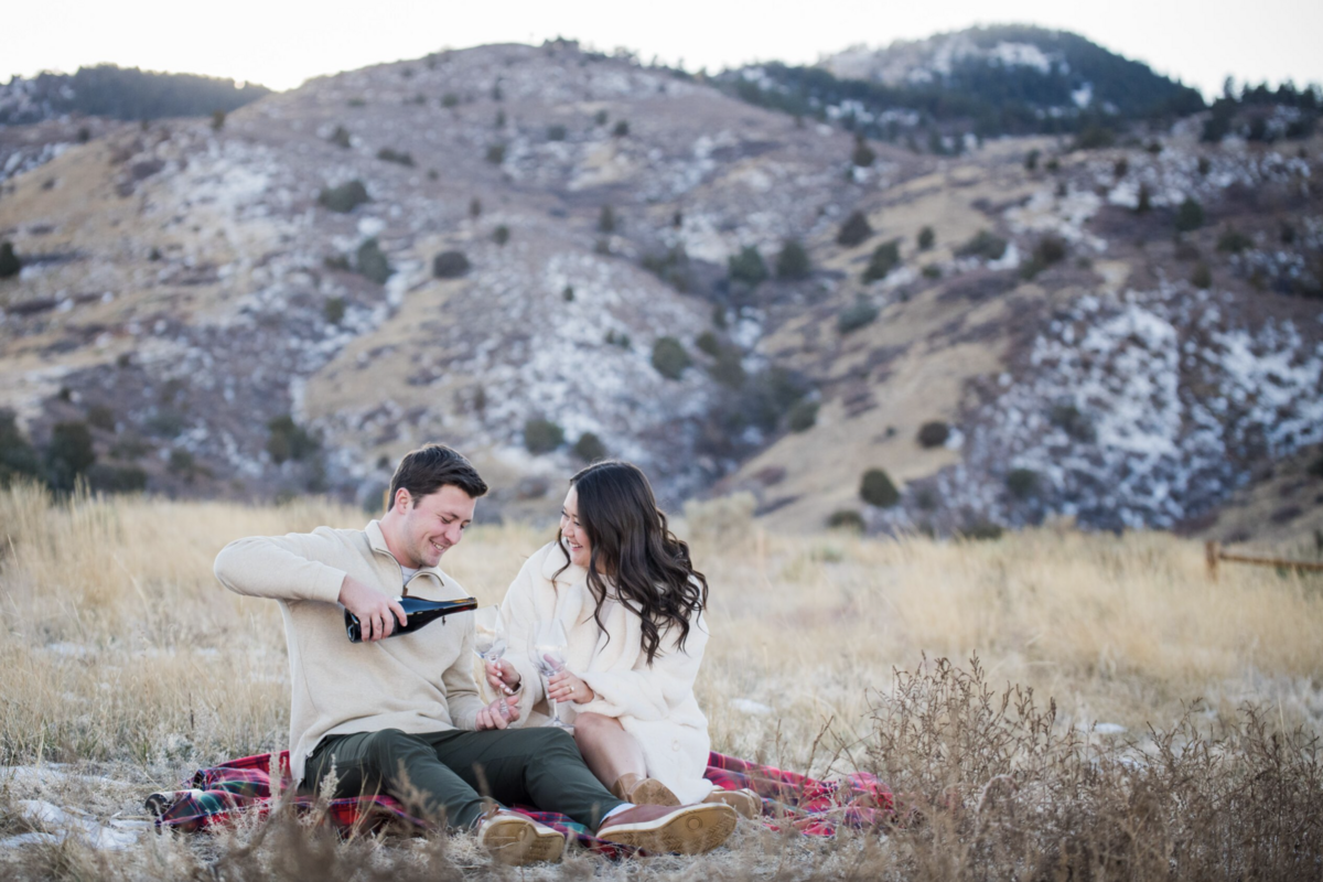 An engaged couple sits on a plaid blanket, and the man pours a bottle of champagne into glasses during their engagement session at Lower Mount Falcon in Colorado.