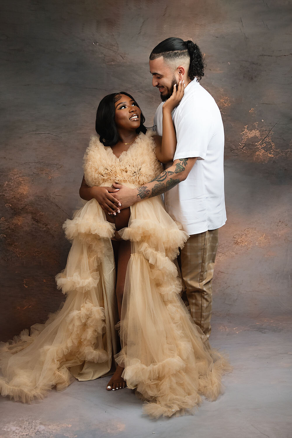 A husband in a white shirt hugs his pregnant wife while they stand in a studio