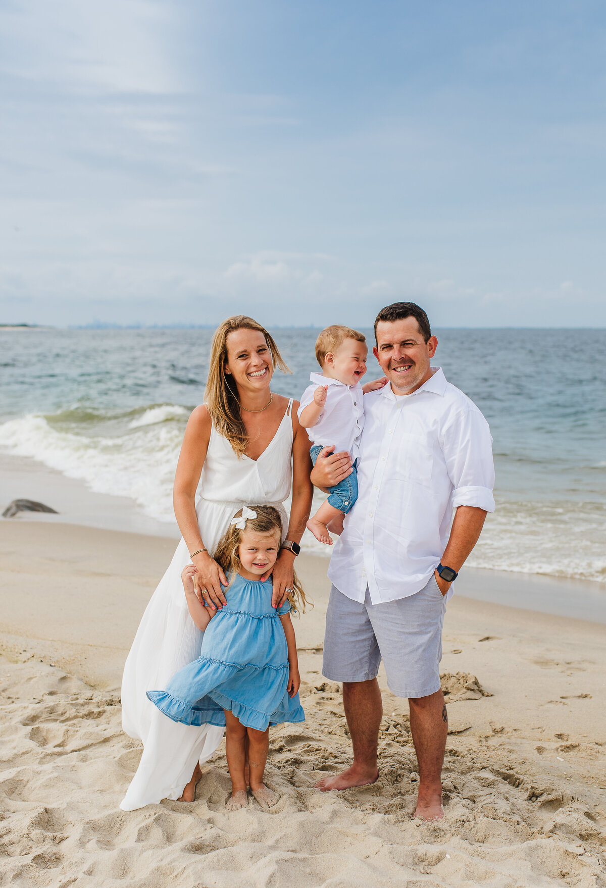 family of four in white and blue standing on the beach in front of the ocean