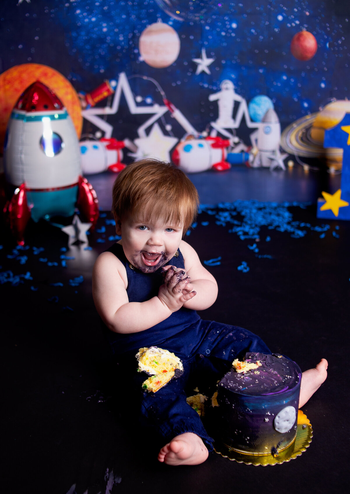 Baby boy wearing blue overalls laughing with cake during cake smash photoshoot in Franklin Tennessee Photography studio, with Franklin Tennessee family photographer