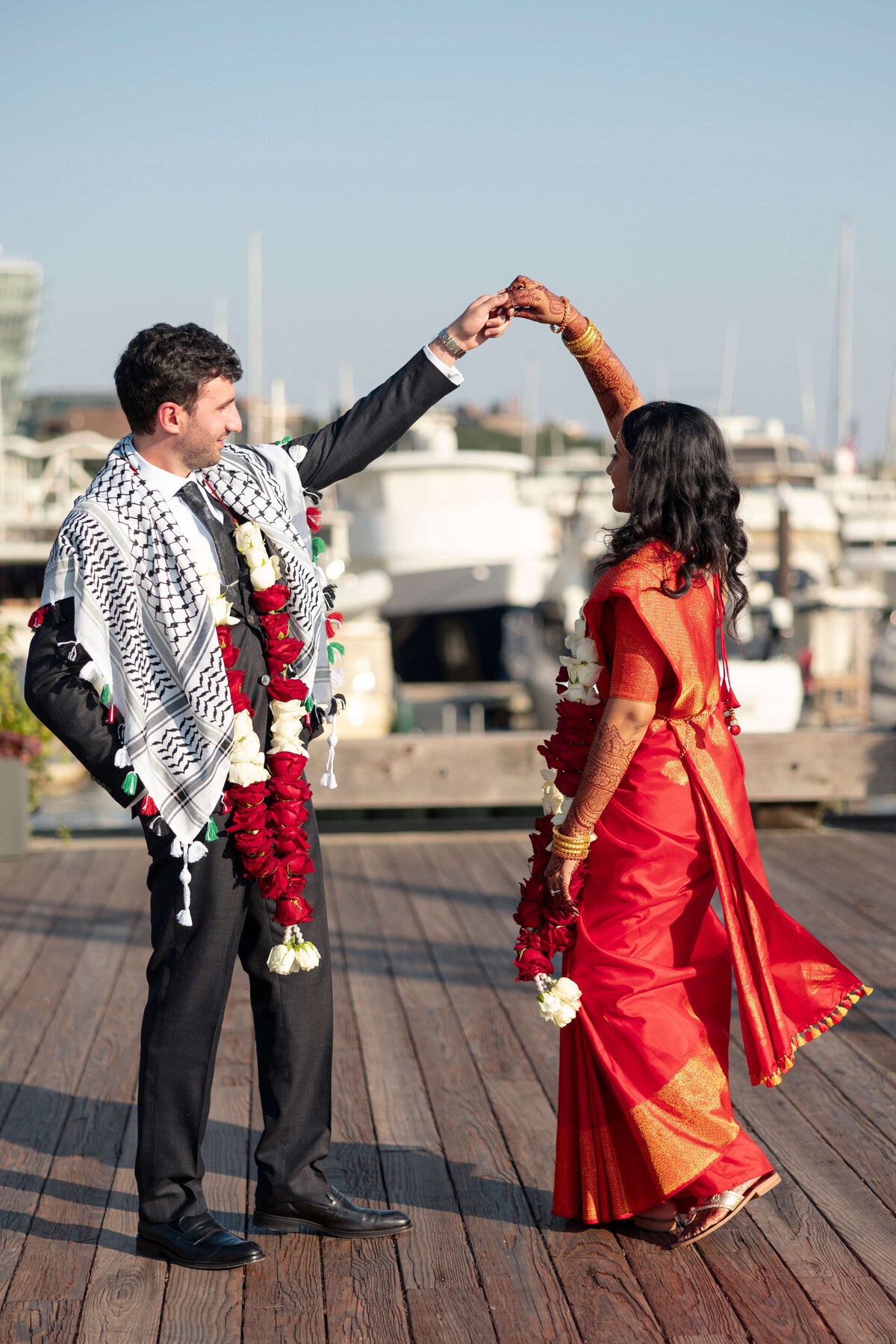 A couple in formal attire dances on a wooden dock. The woman wears a red sari, and the man wears a black suit with a patterned scarf. Both have floral garlands. Boats and a blue sky are in the background.