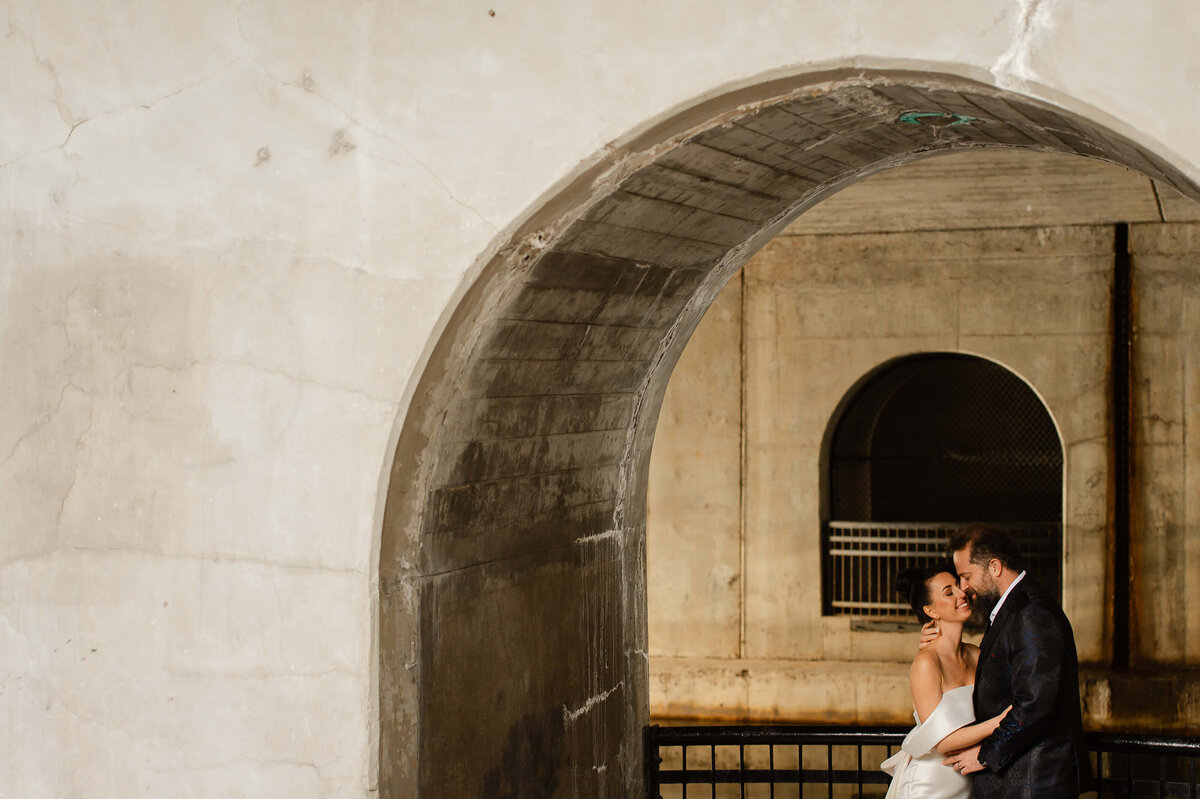 a bride in a white dress and a groom in a blue suit kissing at the Chateau Laurier underpass.  Captured by Ottawa wedding photographer JEMMAN Photography