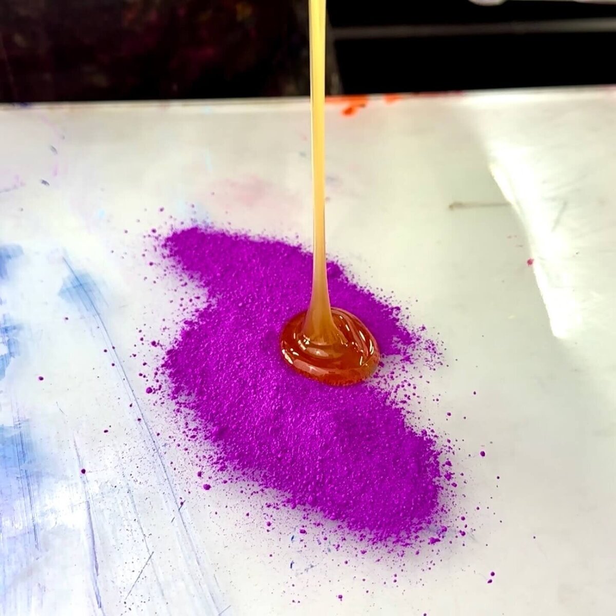 A stream of golden binder is poured into a pile of bright violet pigment powder, marking the start of custom ink mixing.