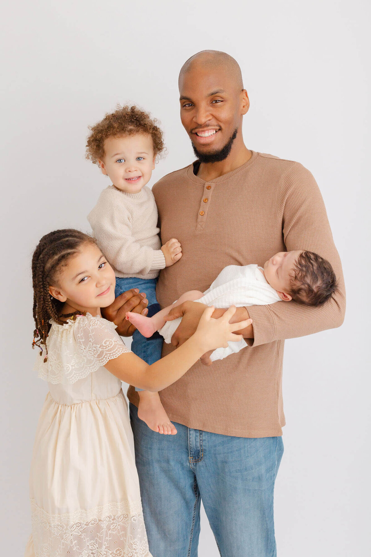 Dad with his three kids. Two older kids and a newborn baby. They are all wearing light, neutral tones and colors at their newborn photo session.