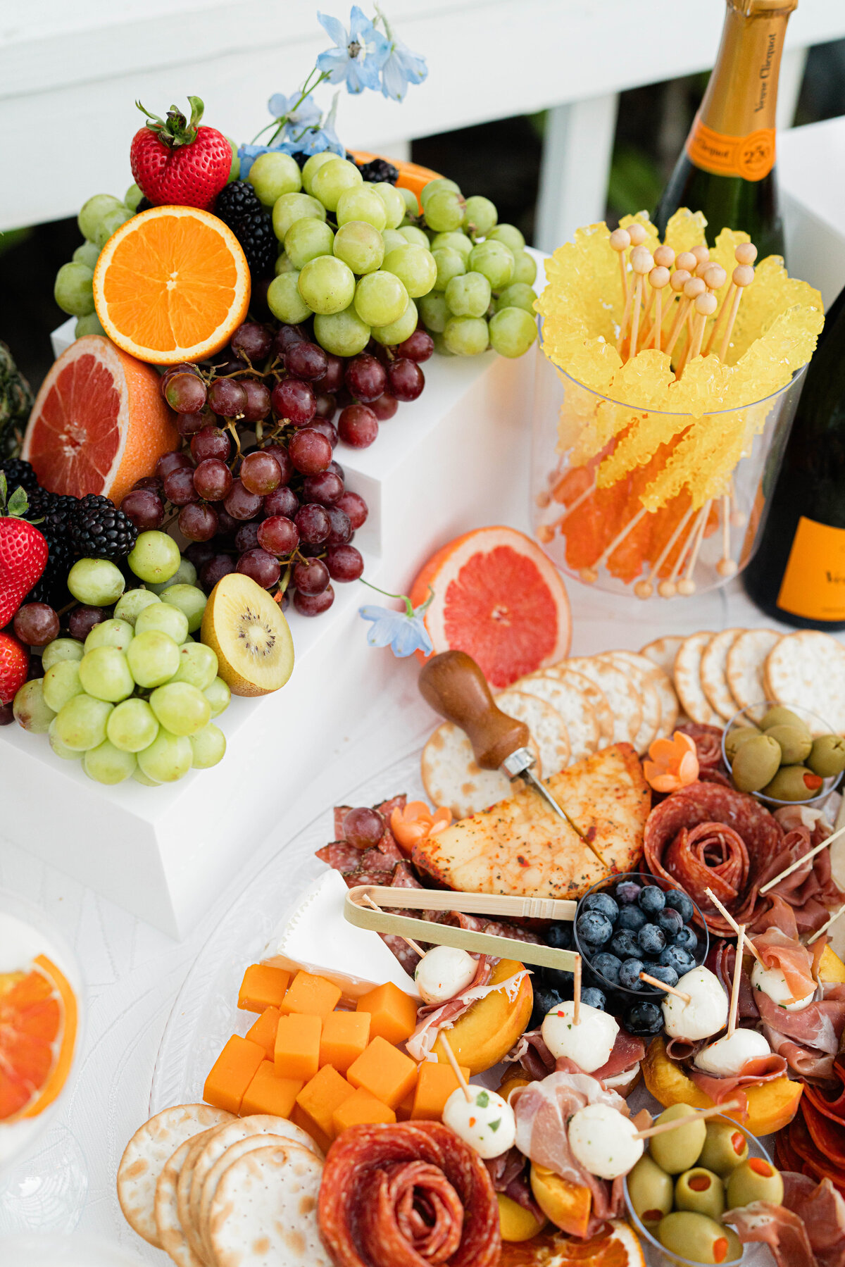 fruit charcuterie of green and red grapes, kiwi halves, strawberries, grapefruit halves, blueberries salami roses and cubed cheddar cheese on white pedestals