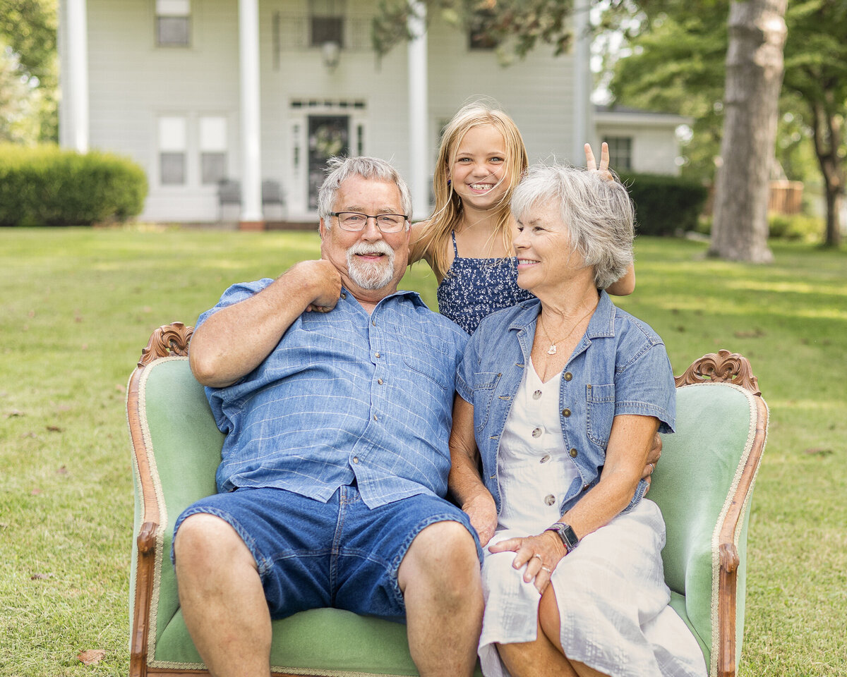 Grandparents on sitting on the vintage green couch in the grass with their family home in the background, with their youngest granddaughter standing behind them, giving grandma bunny ears.  Granddaughter and grandpa are looking at the camera smiling while grandma is looking at grandpa smiling. Grandpa is wearing a light blue and white stripped short sleeve button up shirt, blue jean shorts, and has white short hair and goatee.  Grandma is wearing a white button up dress with a jean jacket over it, with short white hair.Youngest daughter has a dark blue and white tank top style shirt with a blue jean skirt, and has medium length blonde hair.