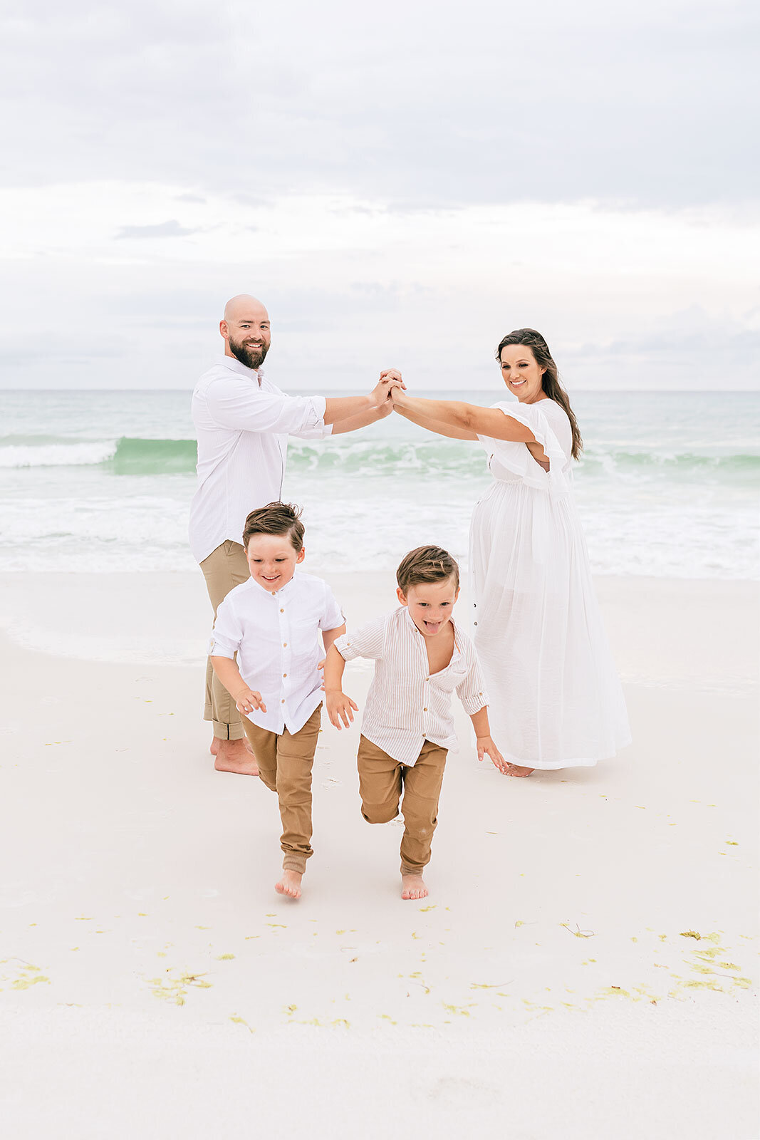 On a sandy beach, a family of four in white and beige outfits enjoys a playful moment. The parents hold hands in the background as the two young boys dash toward the camera, capturing a memory reminiscent of Destin maternity photography by Andrea Krey.