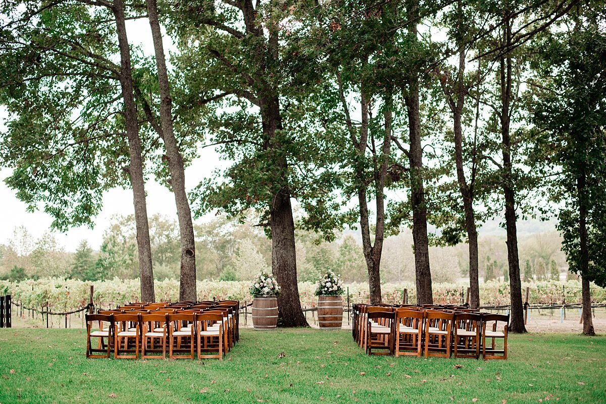 Brown fruitwood chairs are set up in front of tall trees and wine grape vines. On either side of the aisle are two wine barrels topped with two large white and blush floral arrangements.