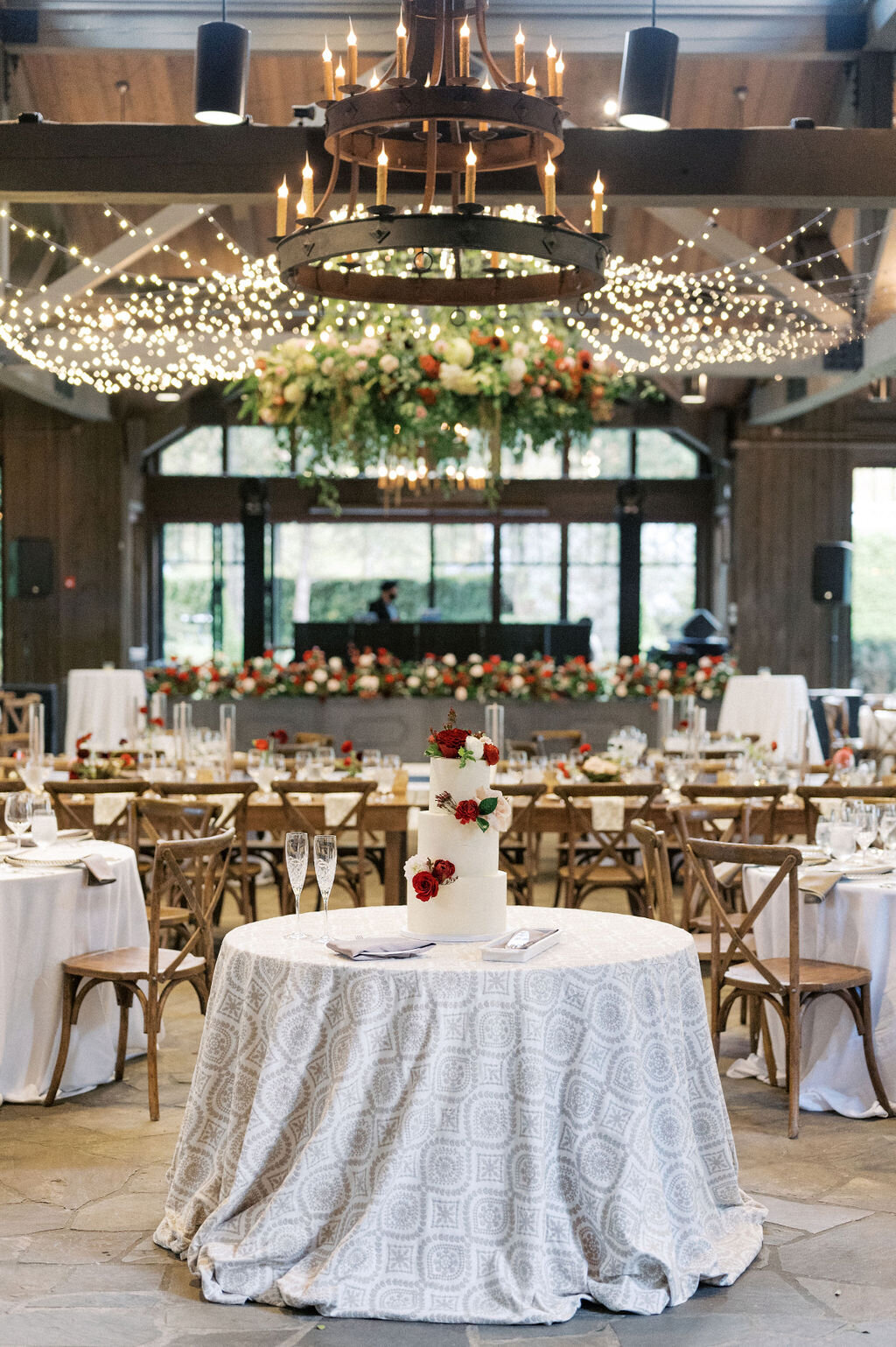 Luxury wedding reception with tiered cake, red floral arrangements and large hanging chandelier installment.