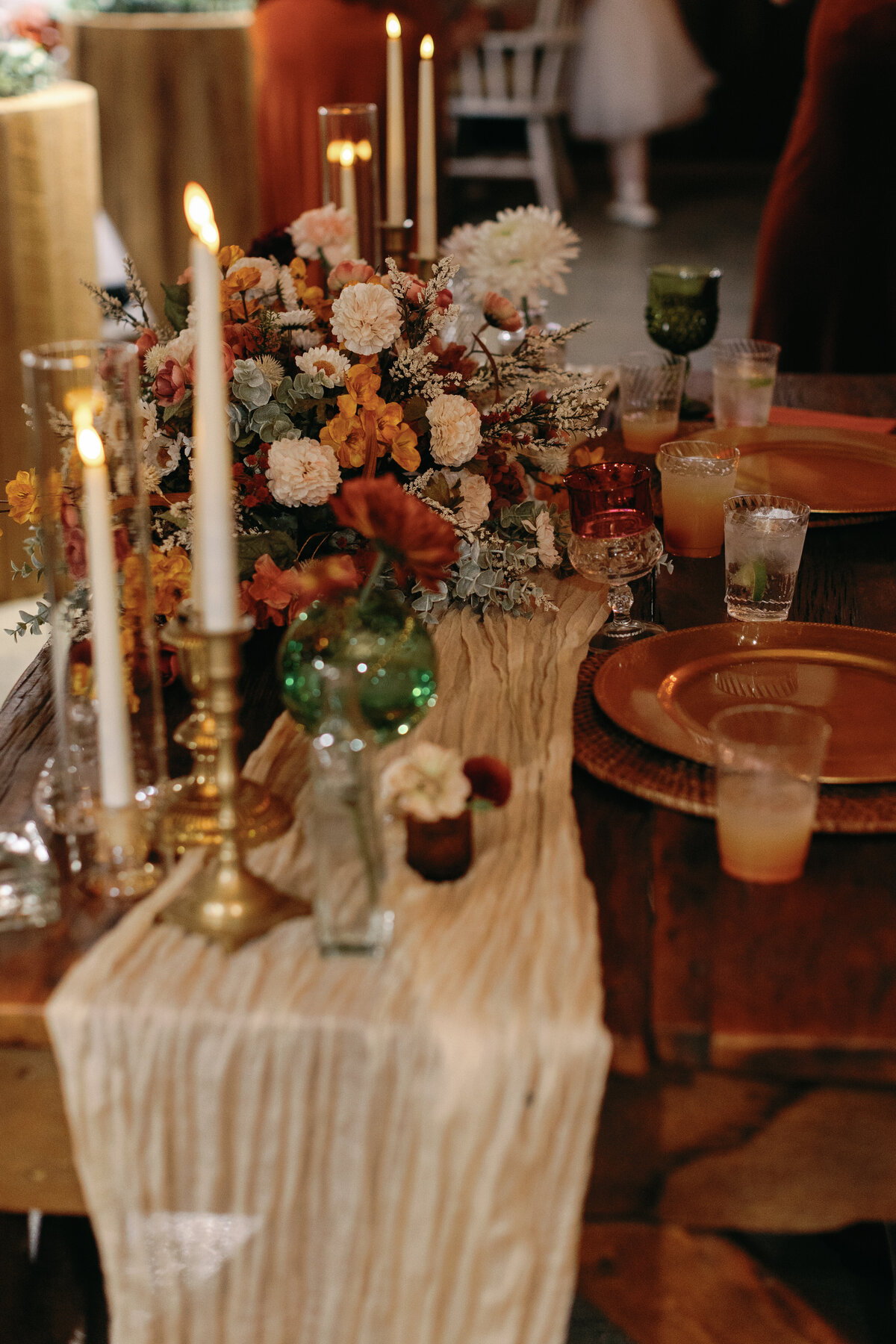 Wedding reception table set with tall gold candlesticks, rust and blush floral centerpieces, and amber glassware inside The Hummingbird Chateau barn.