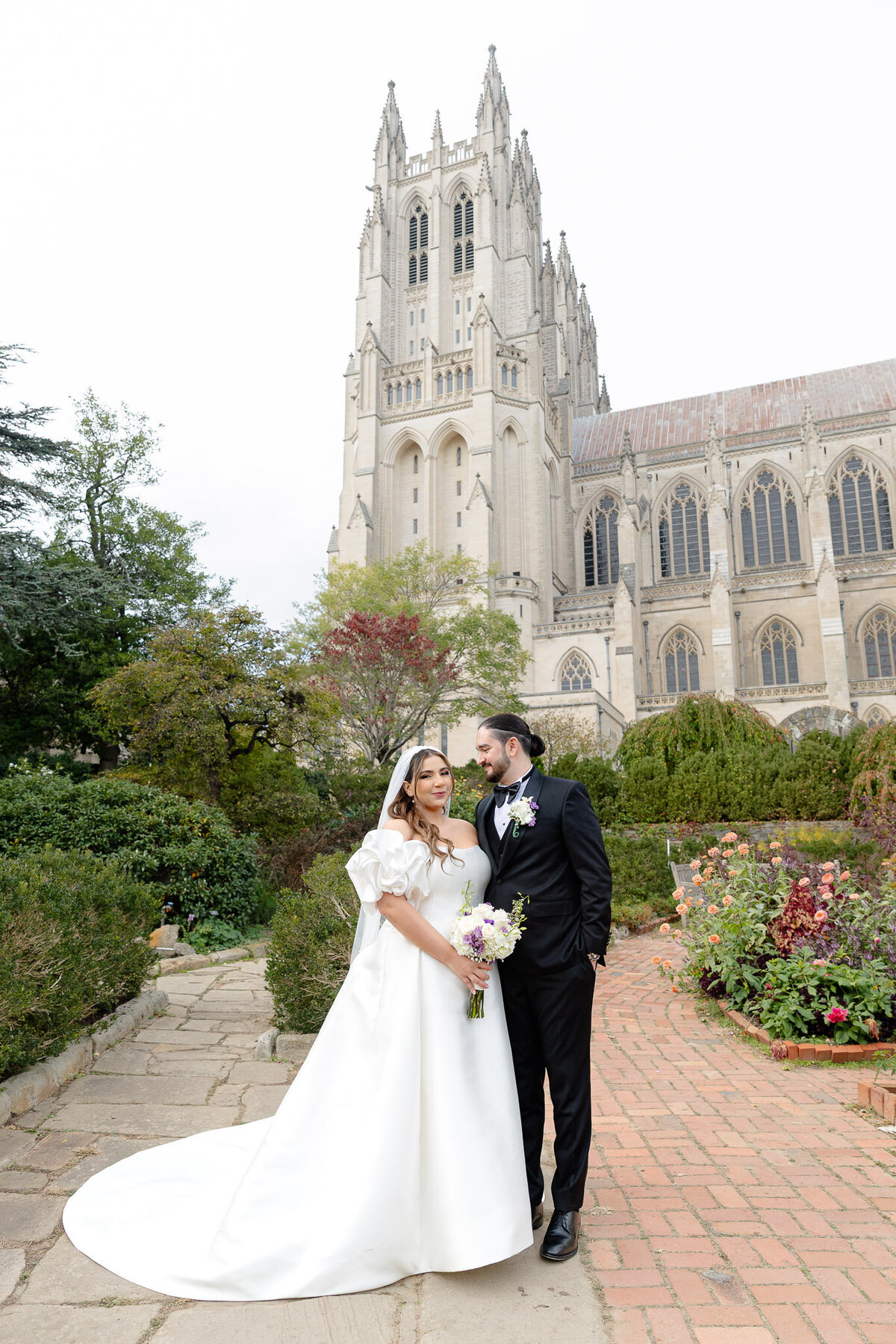 A groom looks at his bride, while she looks at the camera. Behind them is a gorgeous cathedral located in Washington D.C.