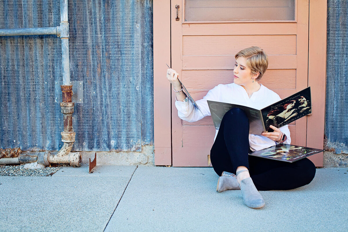 A woman with short blonde hair wearing black pants and a long sleeve, white blouse sitting in front of an orange colored door in a metal wall, looking at a record at Irvine Ranch Park.