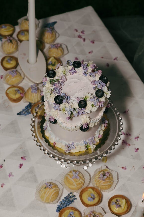A film photo shows a small cake sitting on a clear cake stand, surrounded by spring floral petals and mini pastries scattered on the table. The cake has pale purple frosting with vintage piping in white, pastel pink, and pastel blue, with blueberries and fresh lilac and violet flowers.