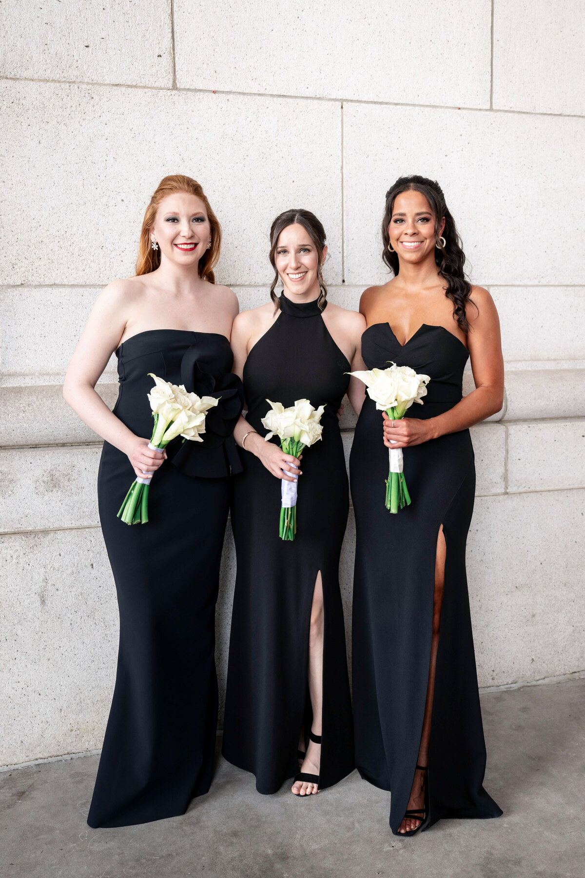 Three women in black dresses stand side by side, each holding a bouquet of white flowers. They are smiling and posing in front of a light stone wall. All dresses have a slit, and they appear to be dressed for a formal occasion.