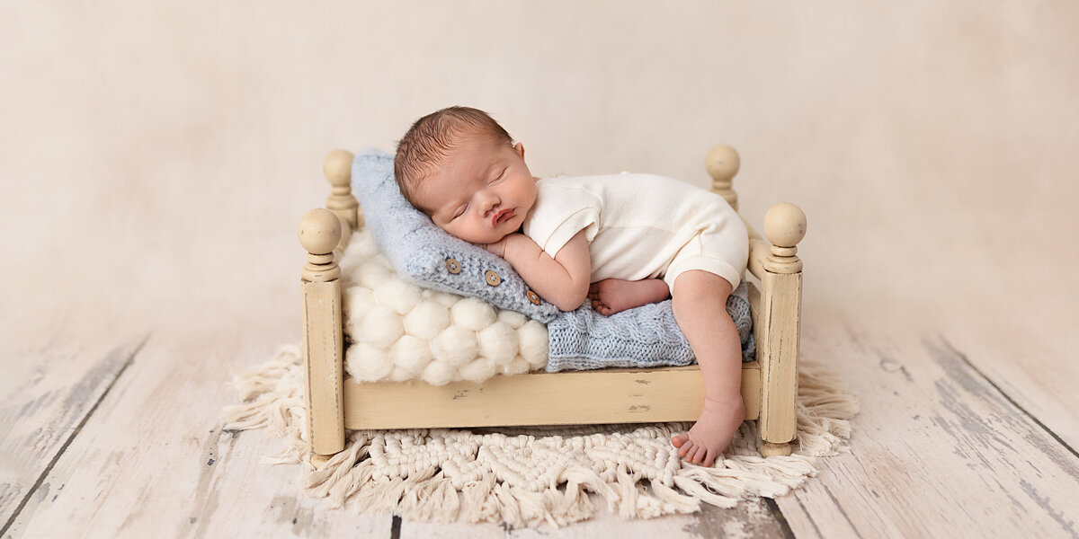 Newborn baby sleeping on a miniature cream bed with a blue knit pillow and textured blanket on a rustic wood floor.
