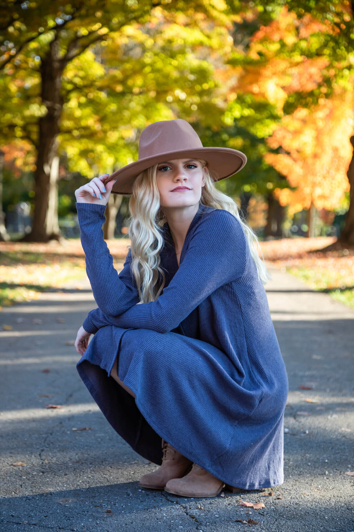 A senior picture of a lovely blonde teen  posing with her arms crossed over her knees in the middle of a Fall leave covered path. Captured by Springfield, MO senior photographer Dynae Levingston.