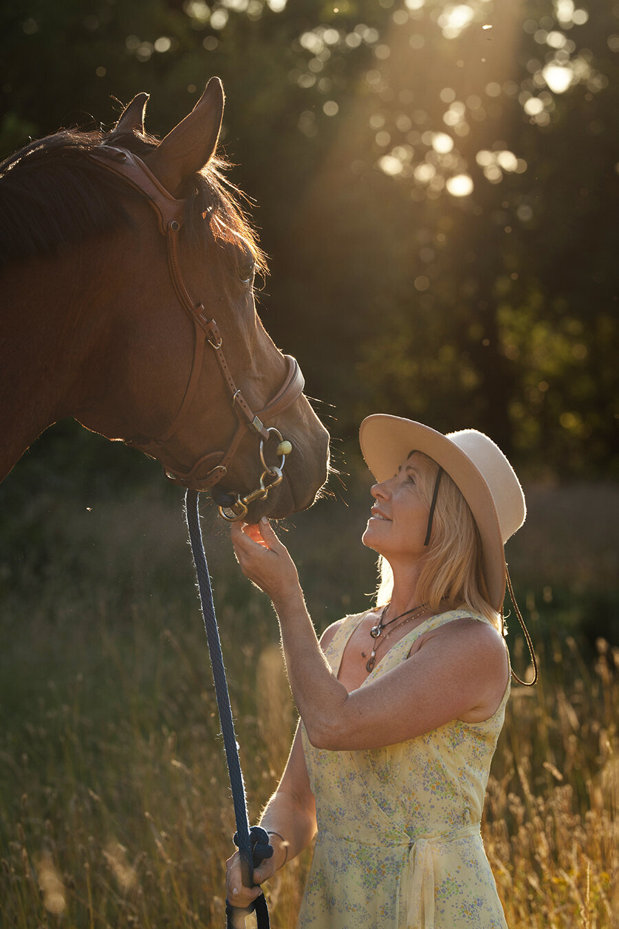 A women in a yellow dress and a cowboy hat is looking at the face of the horse in golden hour