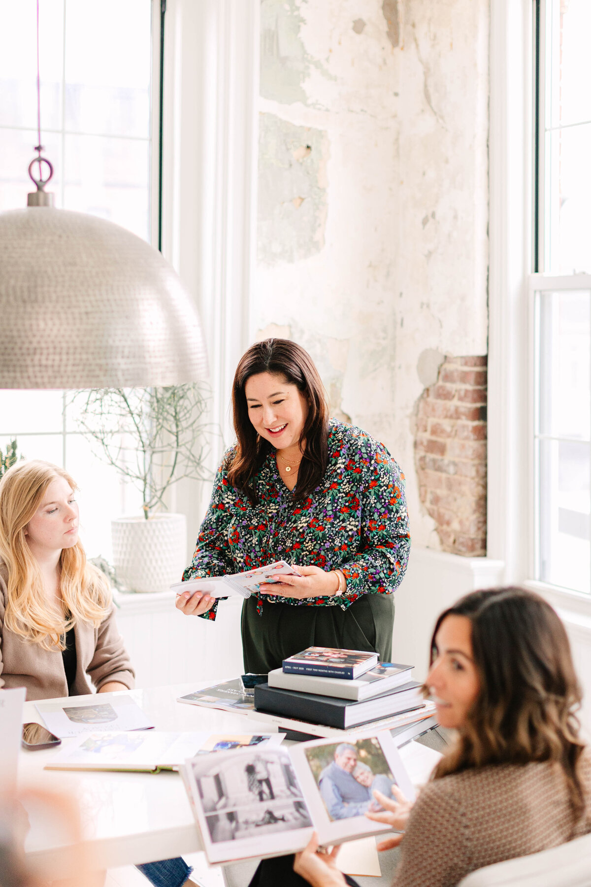 A woman in a floral blouse stands at the head of a table, holding an open book and speaking to a group in a bright, modern workspace. The table is covered with photo albums and design materials, while natural light streams through large windows. The exposed brick and distressed walls add a creative, industrial touch to the setting