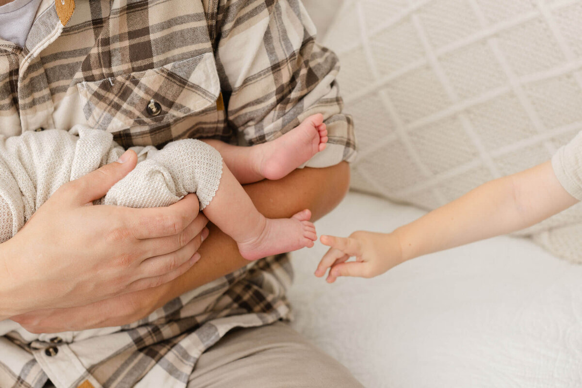 Close-up image of newborn baby feet in dads arms and one little toddler hand is reaching out towards the baby's feet.