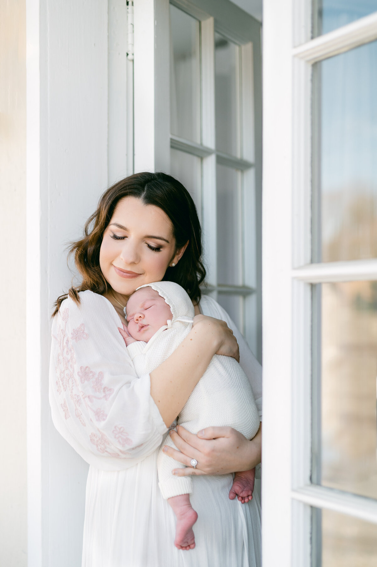 A mom leans against a patio door with her eyes closed, snuggling her newborn baby girl into her chest
