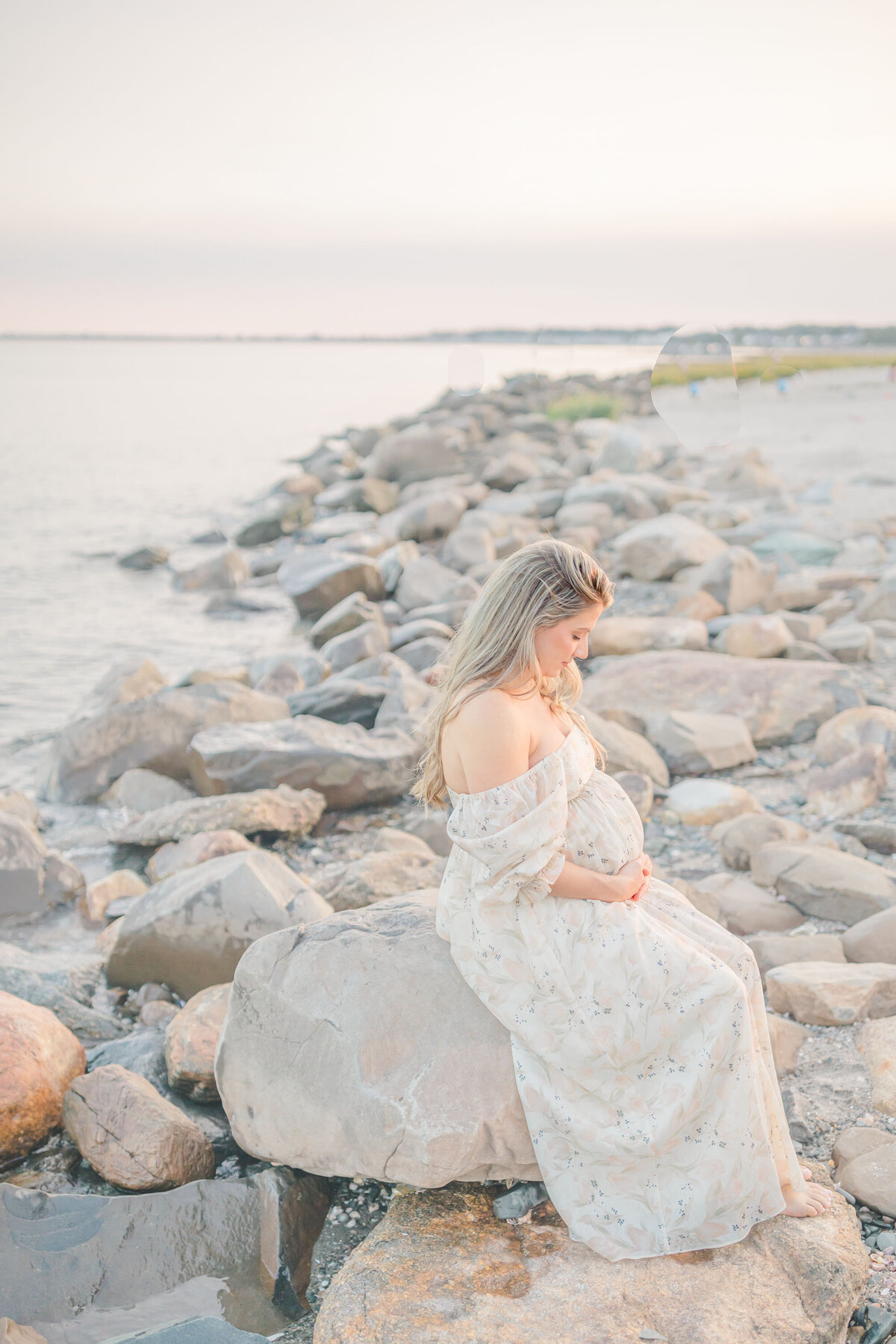 A pregnant mother sits on rocks at the beach, gently resting her hands on her belly. The soft waves of the ocean can be seen in the background, with the warm sunlight casting a peaceful glow over the scene