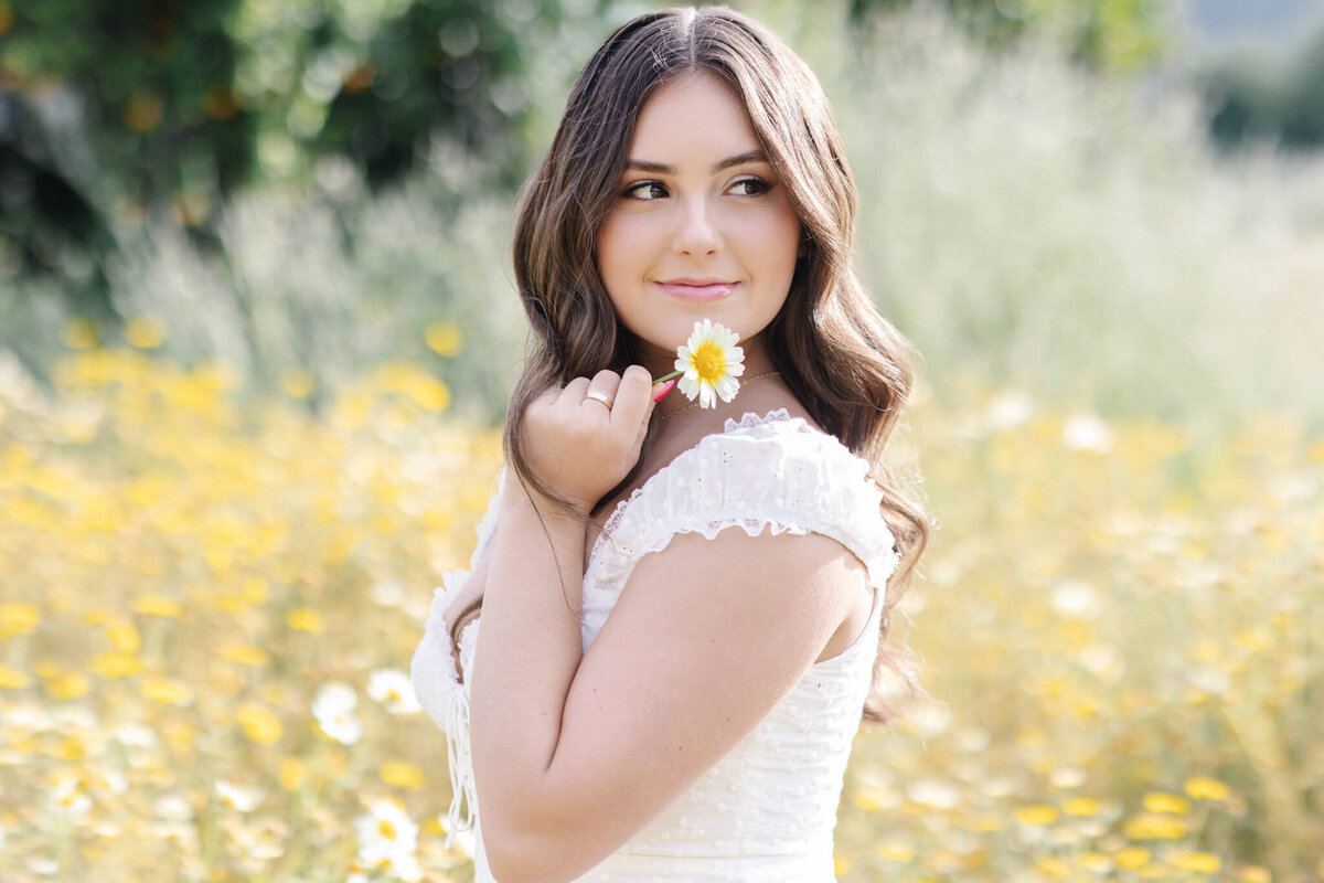 A woman in a white dress stands in a field of yellow and white flowers, holding a daisy to her face. She has long, wavy brown hair and is looking to the side with a gentle smile. The background is blurred, with greenery visible.