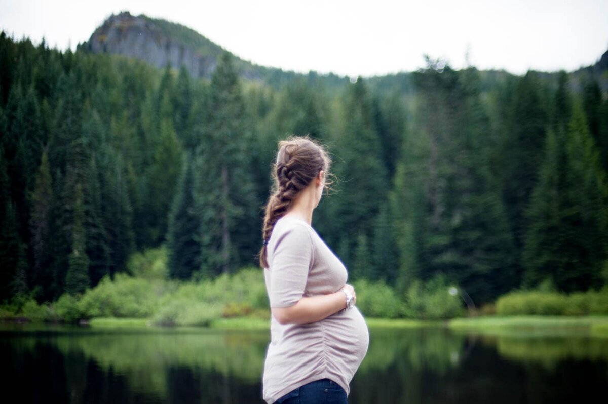 Expecting mother in a tan shirt and braided hair in front of a lake and pine trees.