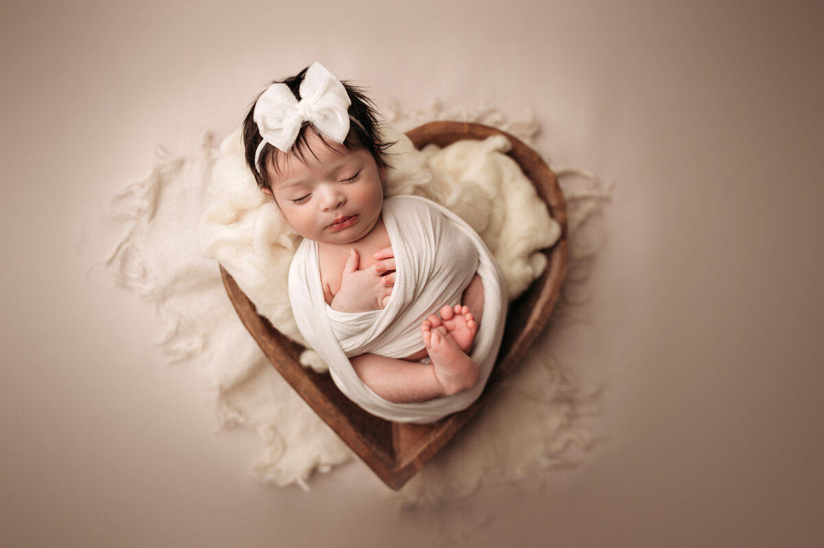 baby girl wearing big whie bow and wrapped in swaddle  posed inside of brown heart bowl prop