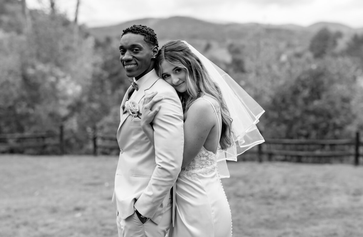 A bride and groom stand closely, smiling at the camera, captured by a documentary photographer for New England weddings. The bride, in a veil and dress, leans on the groom's back. He wears a light-colored suit. They are outdoors with trees and mountains in the background in black and white.